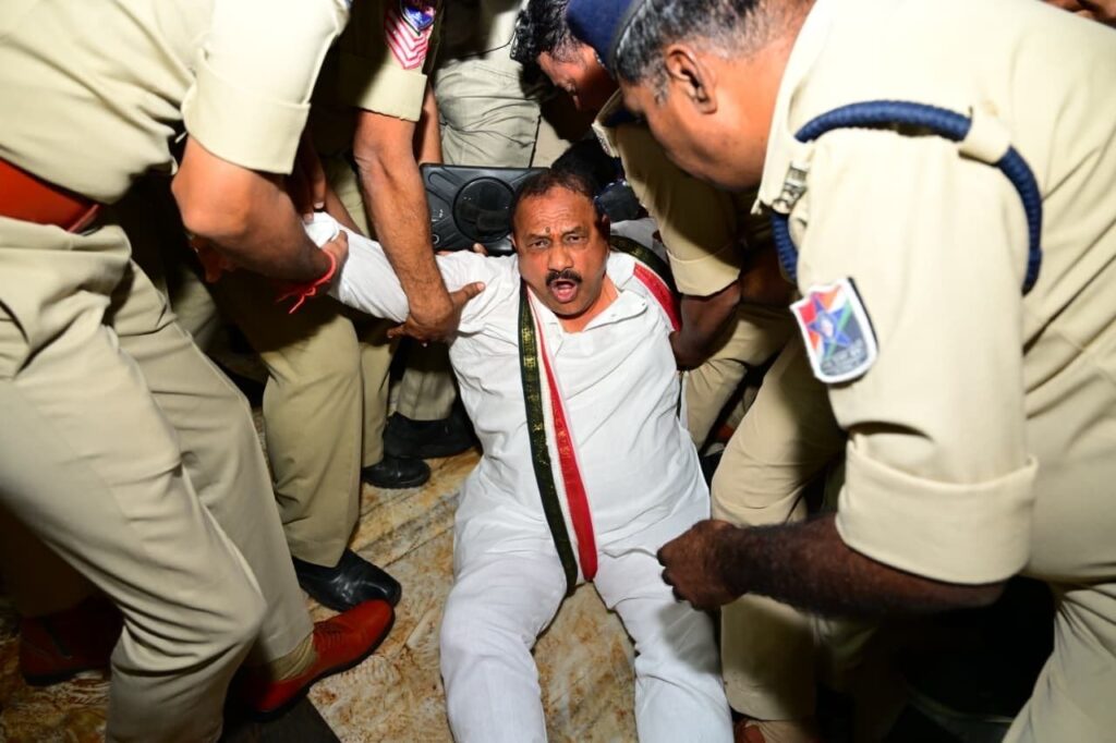 Protest scene with a man in white being restrained by police officers.