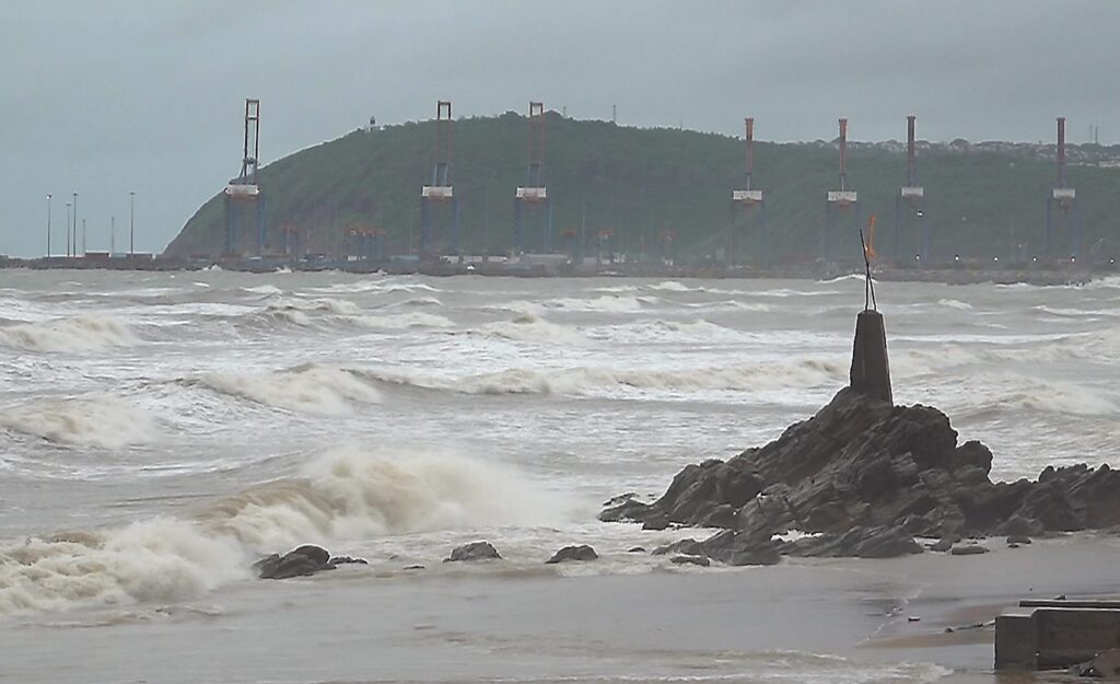 Rough sea with waves crashing against rocks near a coastal breakwater and distant hill with structures.