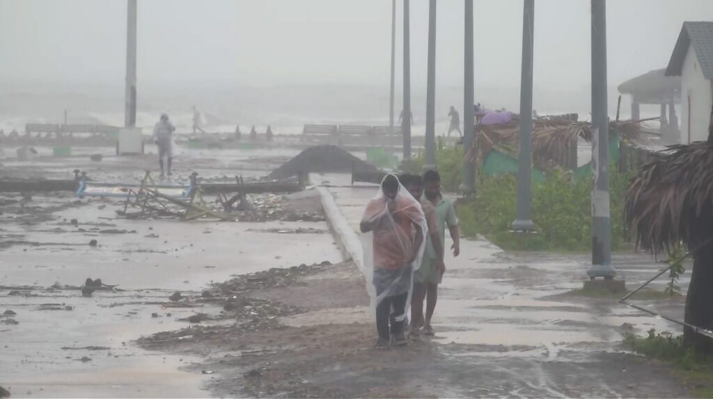 People walk along a flooded, debris-strewn shoreline with makeshift huts and streetlights in the misty,雨y.