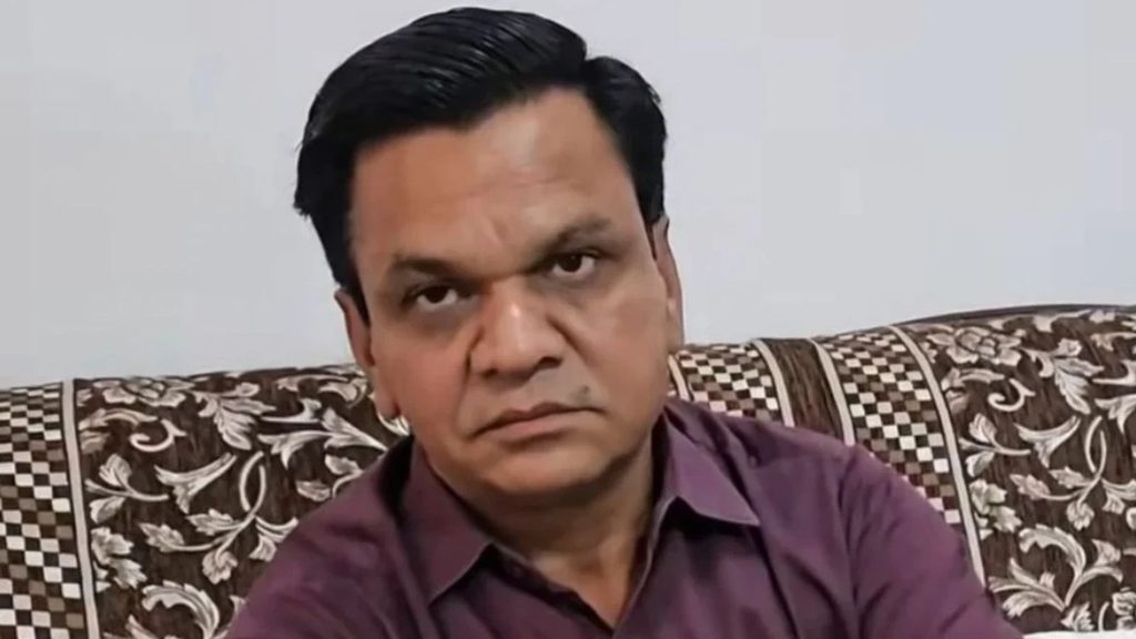 A stern man sits on a patterned sofa in a purple shirt, looking directly at the camera.