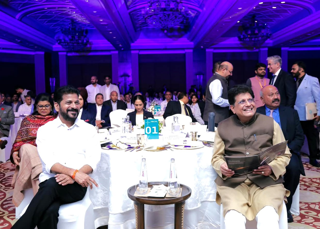 Two men seated at a round table during a formal event at Telangana global summit, with guests mingling in.