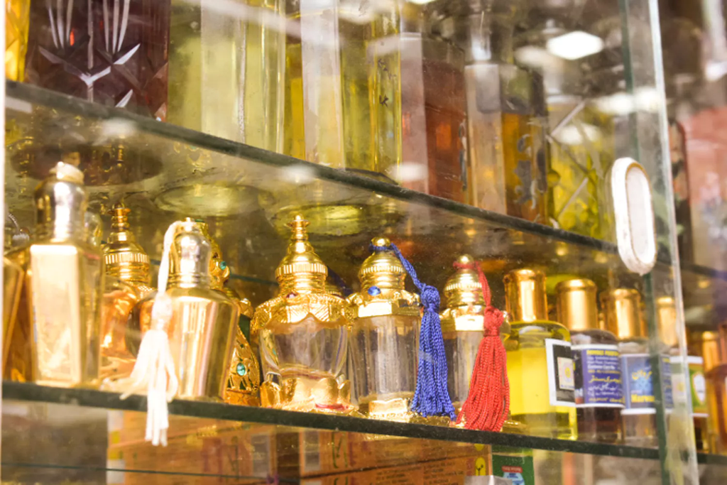 Assorted ornate attar bottles on glass shelves with tassels and golden lids in Hyderabad's historic lanes.