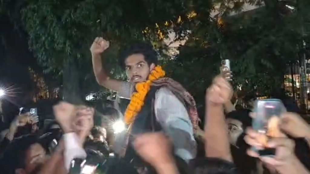Scene of a man wearing a marigold garland raising his fist amid a crowd at night.