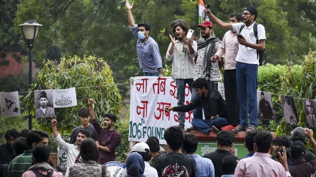 Protesters on a raised platform with banners and photos, raising slogans amid a crowd.