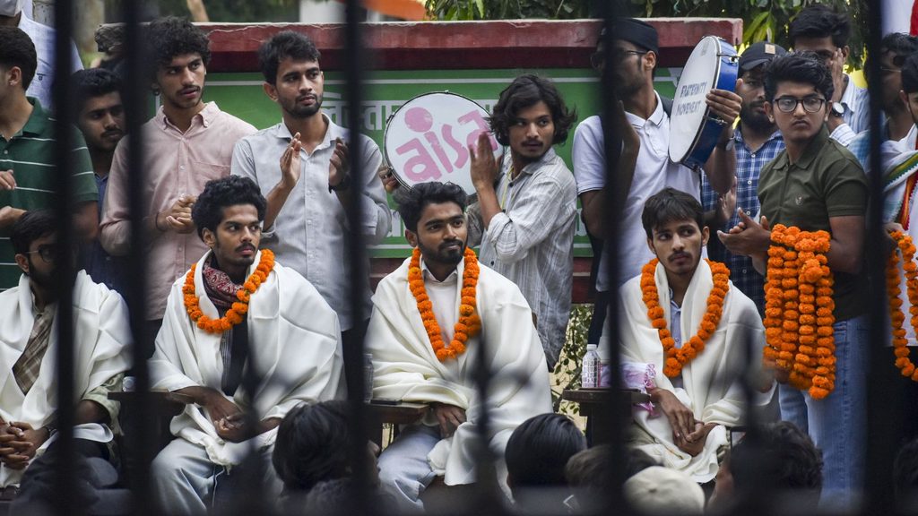 Group of students wearing white robes with orange garlands, gathered behind a barrier during a public学l.